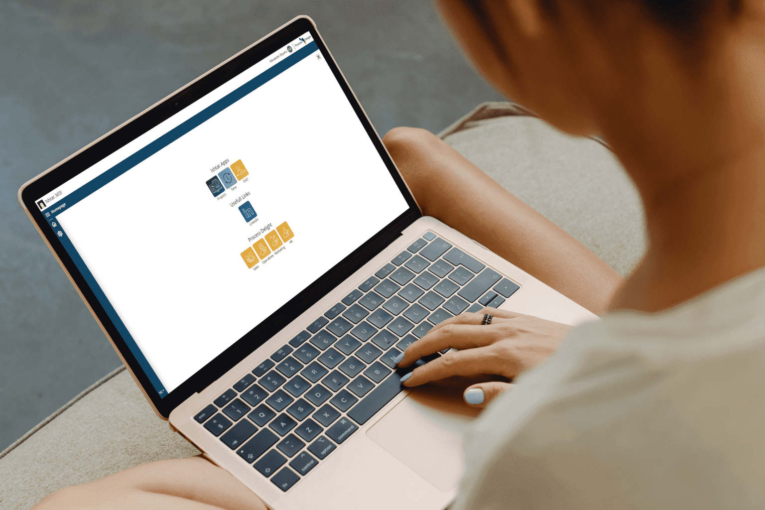 Woman holding a laptop on her legs and sitting in a sofa. On the screen it shows the Ishtar365 platform homescreen with a couple of tiles. There's ishtar.apps tiles; projects, time, OoO (Out of Office). Usefull links tile; Linked in. And Process Delight Tiles; Sales, Operation, Marketing and Hr. Each tile has their own icon.